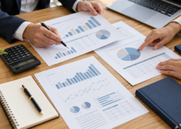 Manos comparando documentos financieros junto a una calculadora y un portátil